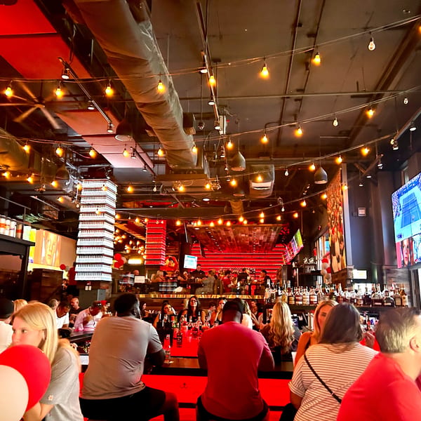 A lively bar interior with red lighting, string lights, and multiple televisions showing sports, filled with patrons.