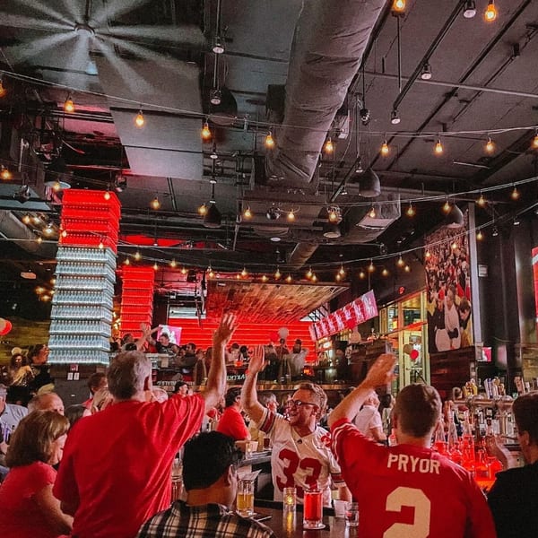 Patrons in red shirts and jerseys cheer at a lively bar with red lighting and string lights.