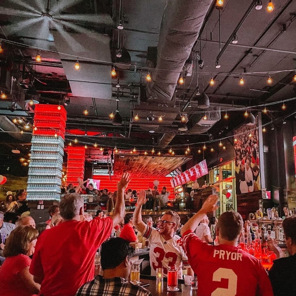 Patrons in red shirts and jerseys cheer at a lively bar with red lighting and string lights.