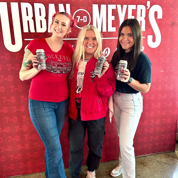 Three smiling women in red and black shirts hold beer cans in front of a red Urban Meyer's wall.