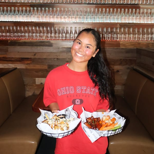 Smiling woman in a red Ohio State shirt holding baskets of fries and chicken wings.