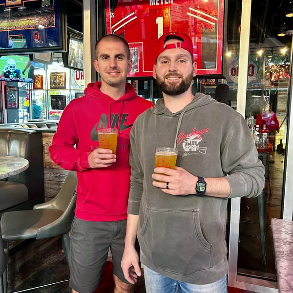 Two men holding beers, smiling in a sports bar with framed jerseys and TVs in the background.