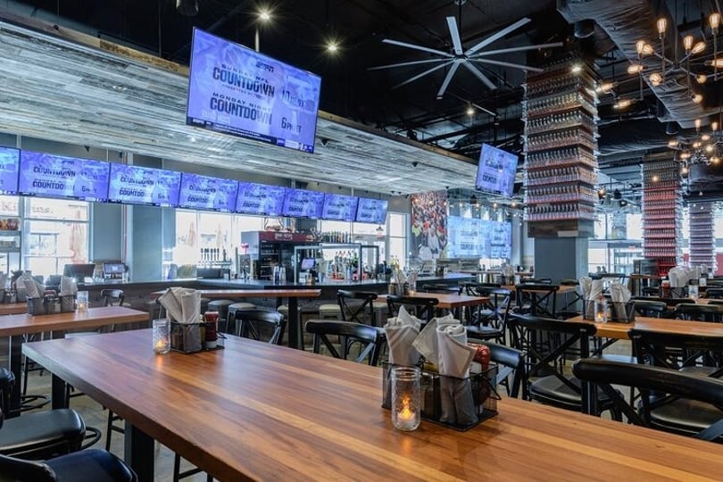 Spacious sports bar interior with multiple large screens, wooden tables, and a long bar area.
