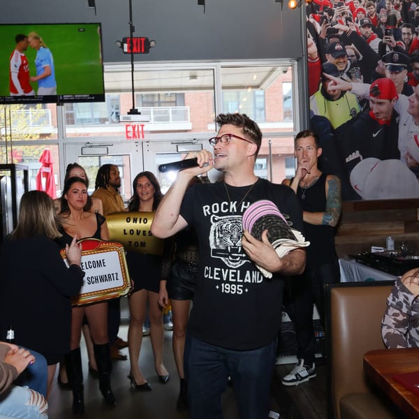 A man in glasses and a black t-shirt sings into a microphone, surrounded by people in a restaurant.