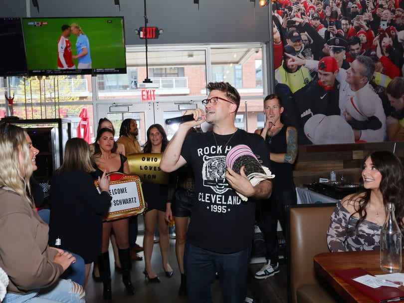 A man in glasses and a black t-shirt sings into a microphone, surrounded by people in a restaurant.