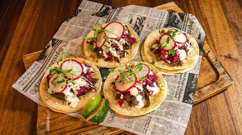 Four gourmet tacos with meat, radish, and microgreens on newspaper-lined wooden board.