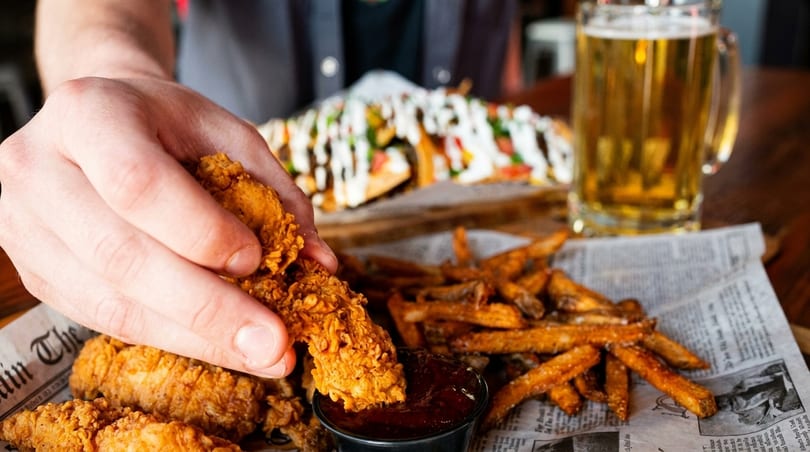 Hand dipping crispy chicken tender into sauce, with fries, nachos, and beer on a table.