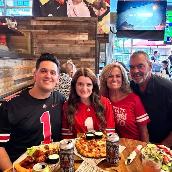 Four smiling people in Ohio State jerseys at Urban Meyer's Pint House, with pizza, nachos, and beer.