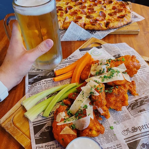 A hand holds a beer mug next to buffalo wings, celery, carrots, and a pepperoni pizza.