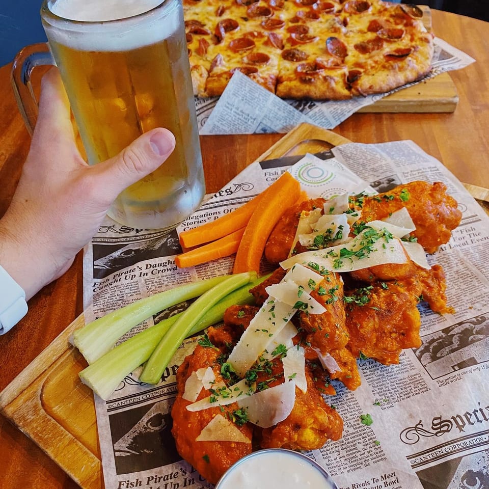 A hand holds a beer mug next to buffalo wings, celery, carrots, and a pepperoni pizza.