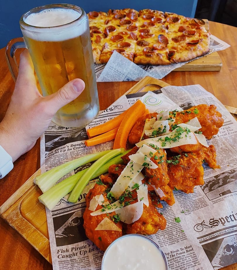 A hand holds a beer mug next to buffalo wings, celery, carrots, and a pepperoni pizza.