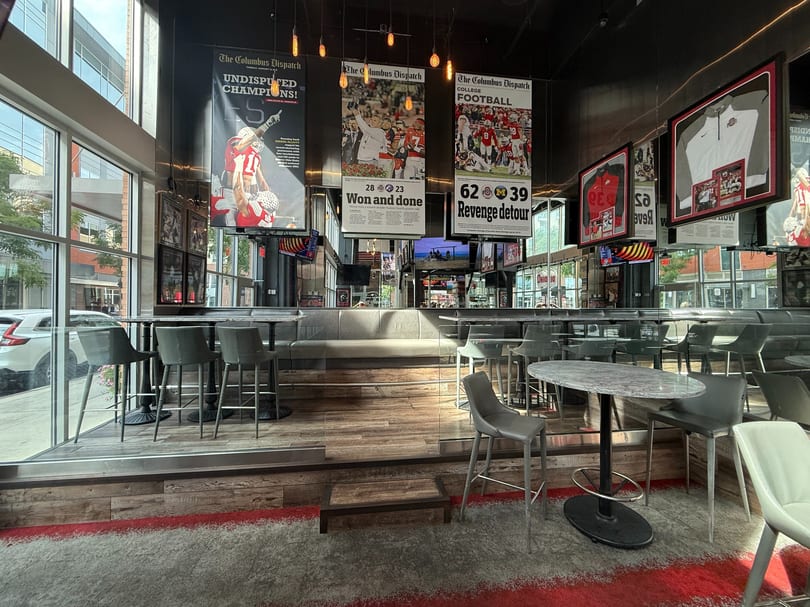 Interior of a sports bar with high-top tables, bar stools, and large sports-themed newspaper clippings.