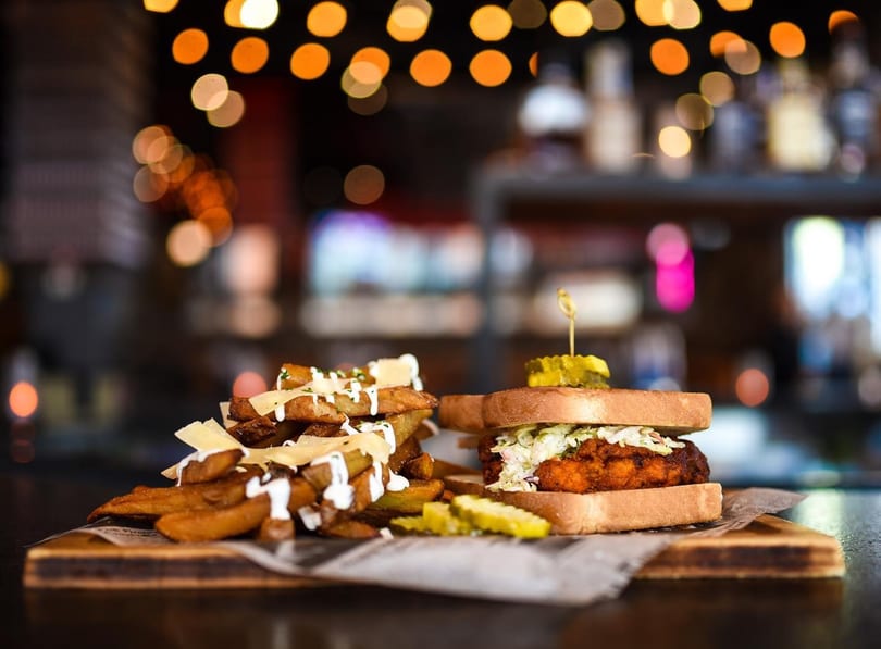 A fried chicken sandwich with coleslaw and pickles, alongside a pile of loaded fries.