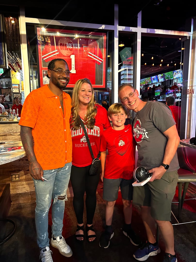 A man in an orange shirt poses with a family of three, all smiling in a restaurant.