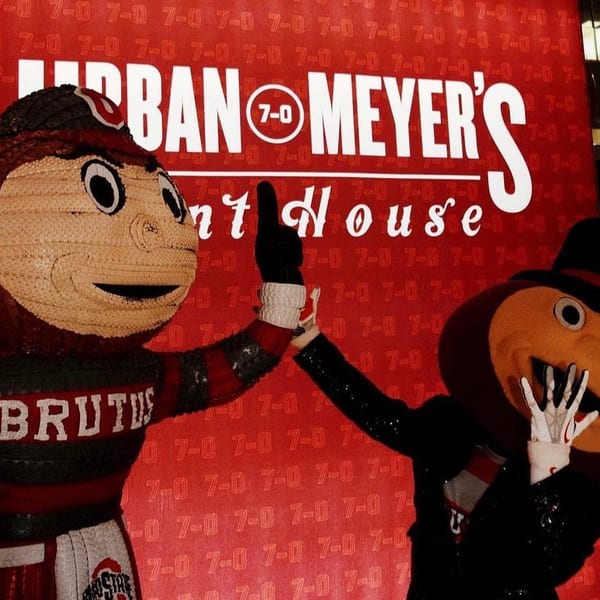 Brutus Buckeye and another mascot high-fiving in front of a red sign for Urban Meyer's Pint House.