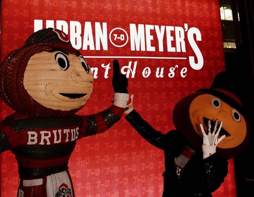 Brutus Buckeye and another mascot high-fiving in front of a red sign for Urban Meyer's Pint House.