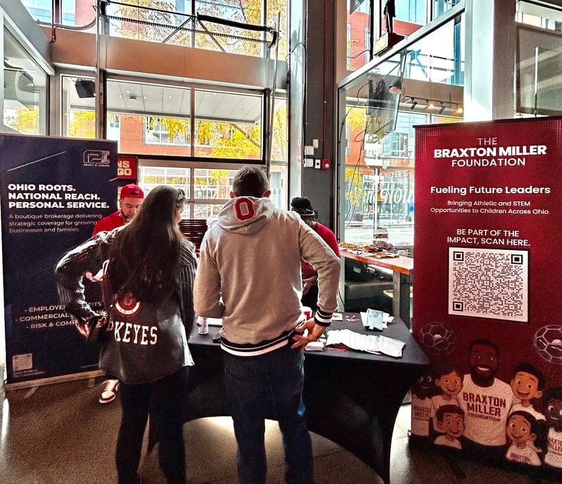 People gather around tables with promotional banners for Ohio Roots and The Braxton Miller Foundation.
